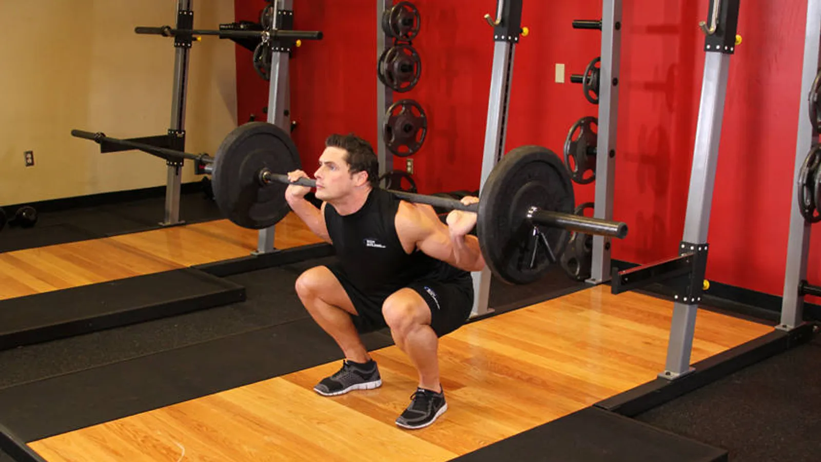 Man at the bottom of a barbell back squat in a squat rack, thighs below parallel, in a gym with red walls