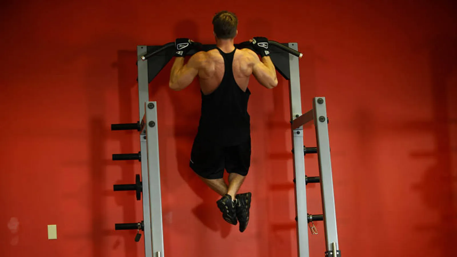 Man at the top of a pull-up, chin above the bar, seen from behind in a gym